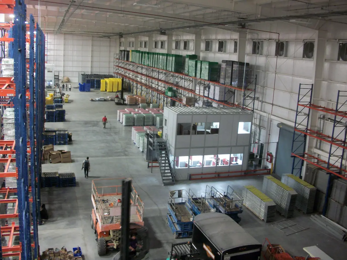 Interior amplio de almacén con estantes metálicos, pallets, dos personas caminando y cabina de control.