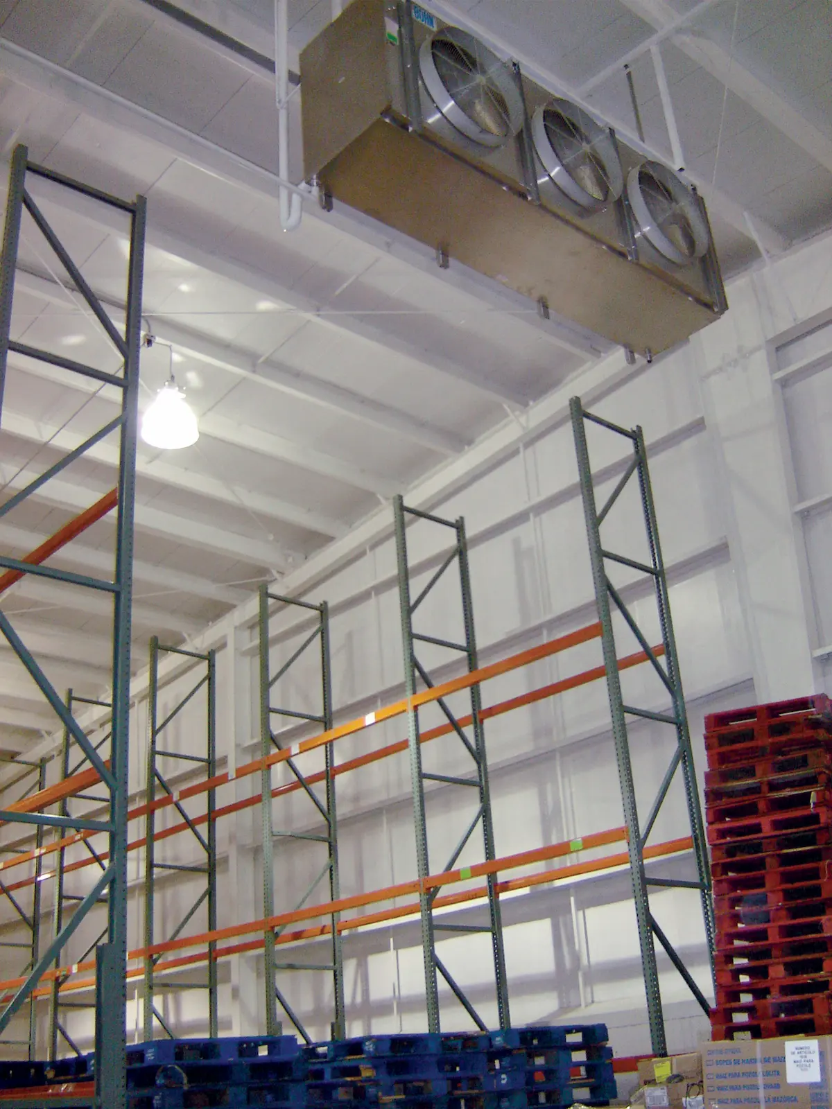 Estanterías industriales vacías y montacargas en un almacén con pallets apilados y ventiladores de techo.