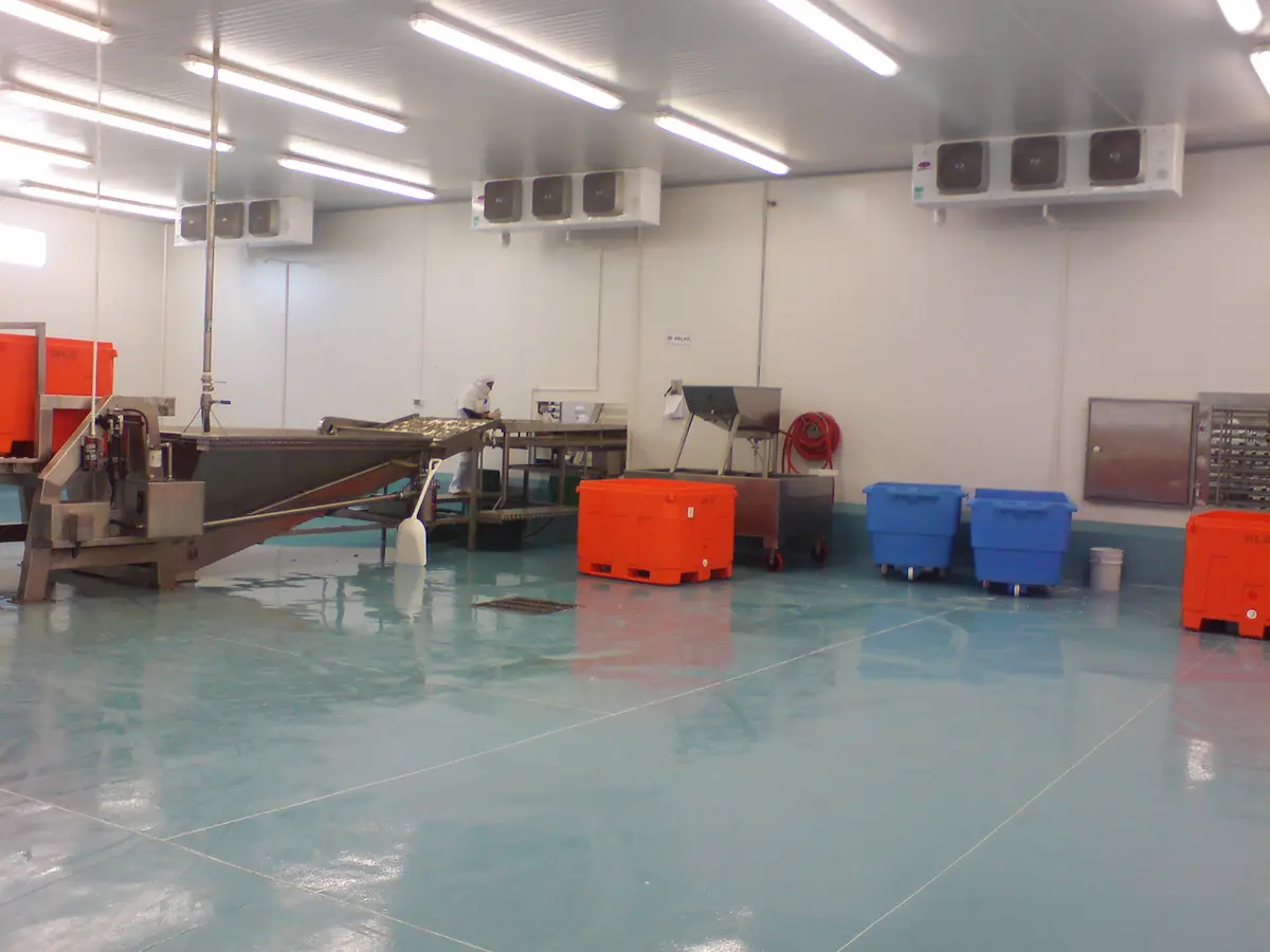 Interior de sala limpia industrial con máquinas y contenedores naranjas y azules sobre piso azul.