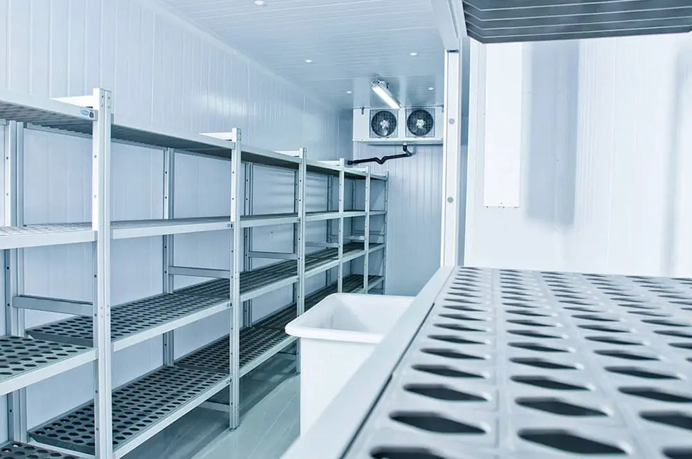 Estanterías metálicas vacías en una cámara frigorífica blanca con sistema de ventilación en el fondo.