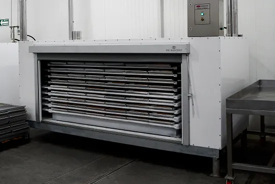 Congelador de placas industrial blanco con bandejas metálicas en un piso industrial.