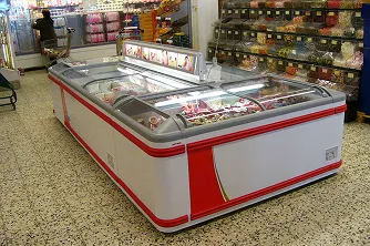 Vitrina congeladora blanca y roja en supermercado con productos congelados dentro y estantes al fondo.