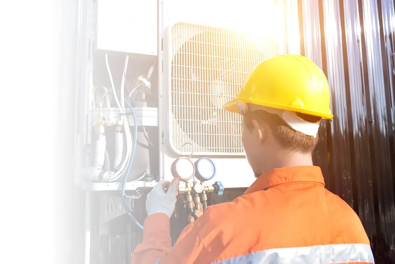 Técnico en casco amarillo y uniforme naranja ajustando medidores en un aire acondicionado externo.