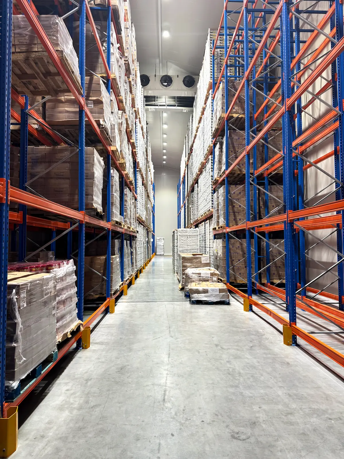 Pasillo de almacén con estanterías azules y naranjas llenas de cajas y palets apilados.