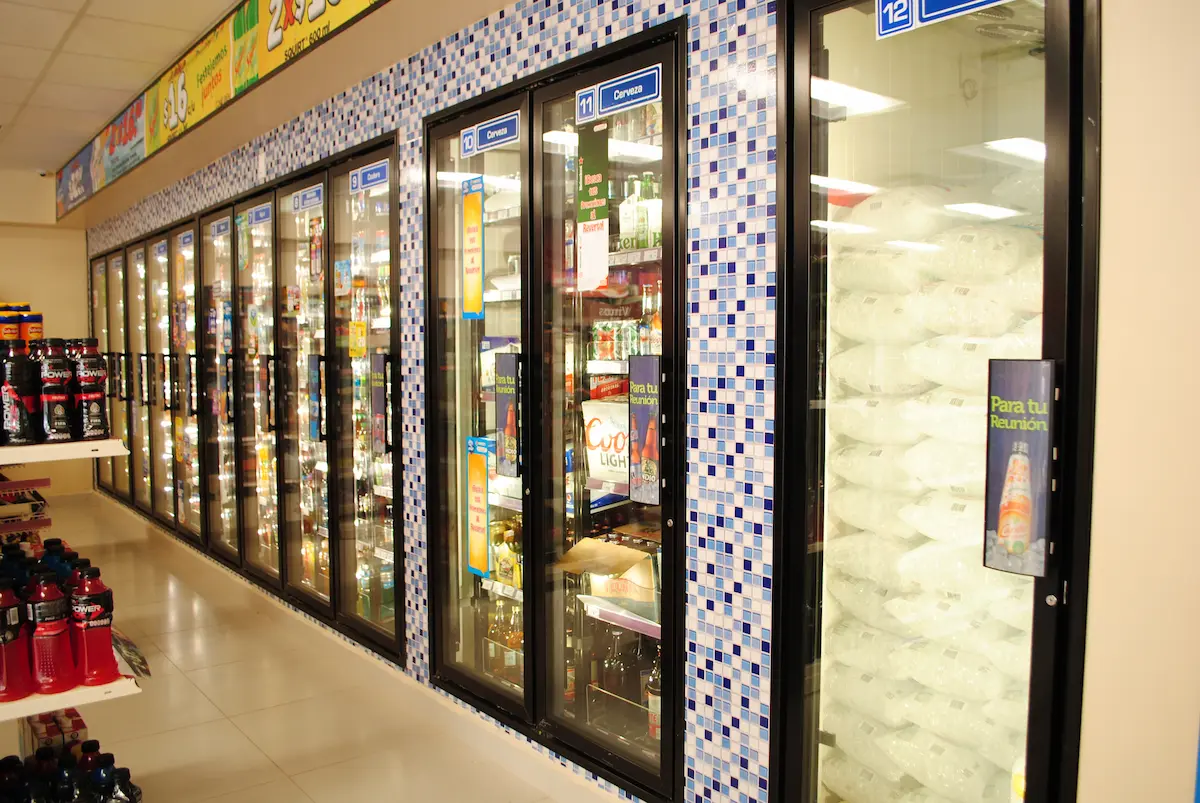 Pasillo de refrigeradores con bebidas y bolsas de hielo en tienda, decorado con mosaicos azules.