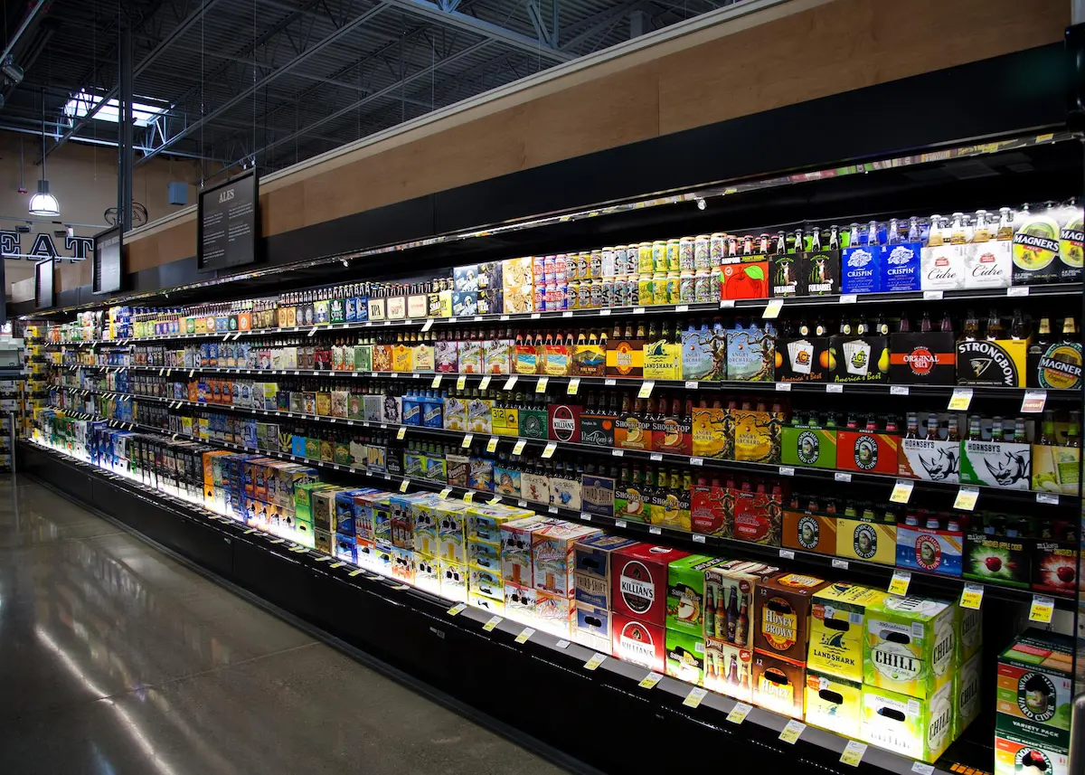 Estante largo con varias marcas de cervezas y sidras en cajas y botellas en un supermercado.