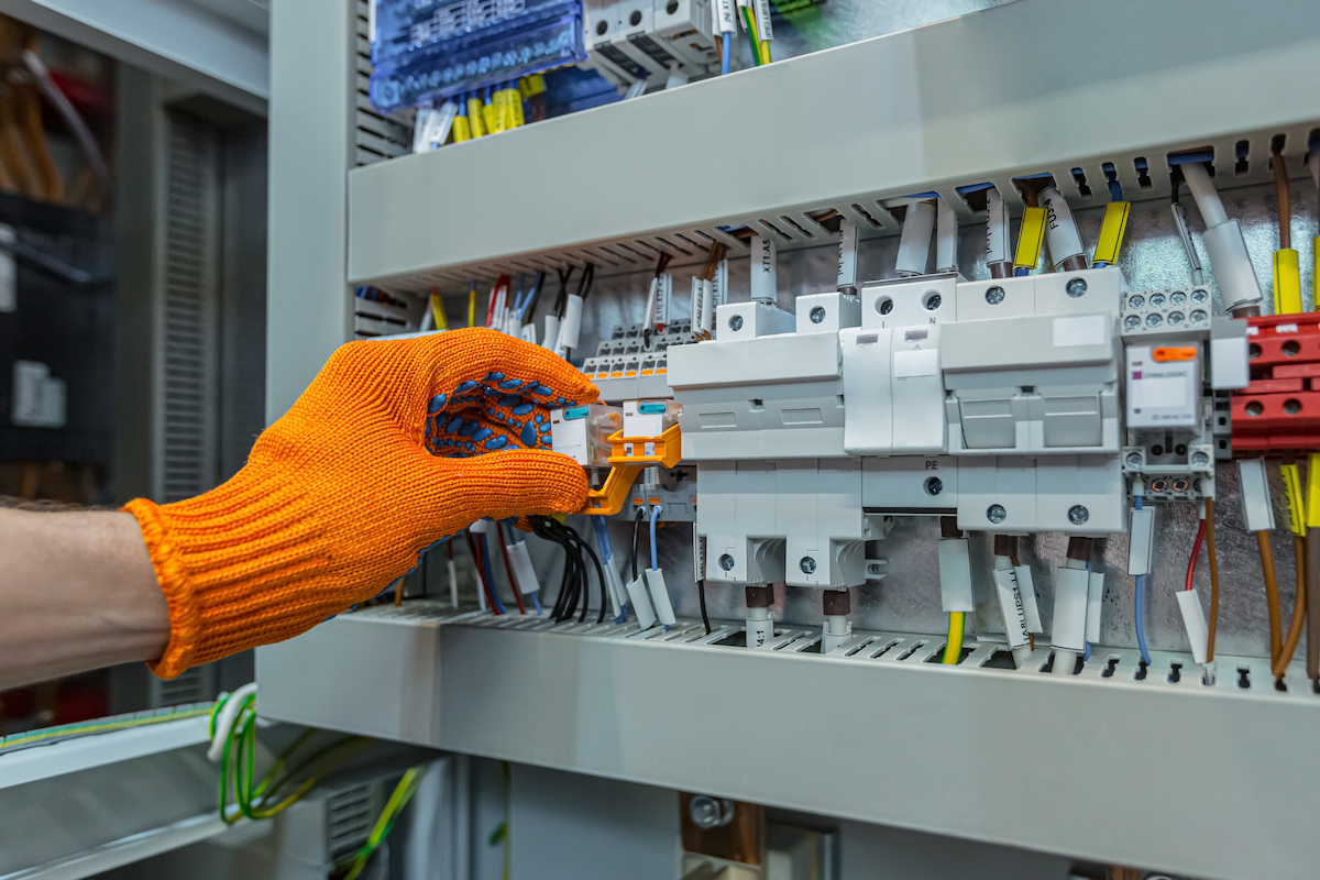 Mano con guante naranja ajustando interruptor en panel eléctrico con cables y componentes.