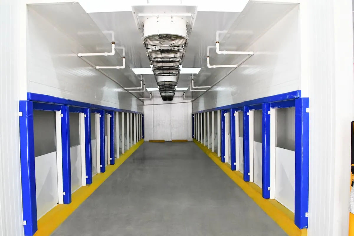 Pasillo largo con múltiples compartimentos blancos protegidos por bordes azules y ventiladores en el techo.