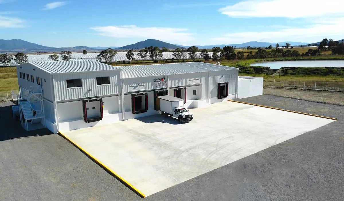 Edificio industrial blanco con camión de carga en área amplia al aire libre con montañas al fondo.