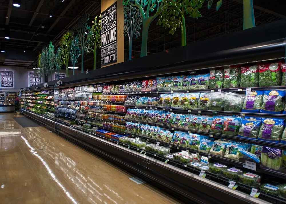 Pasillo largo de supermercado con estantes llenos de productos frescos y verduras en bolsas.