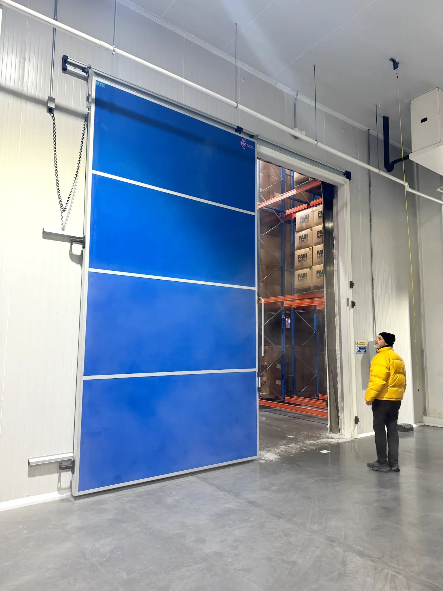 Un hombre con chaqueta amarilla frente a una puerta azul corredera en un almacén.