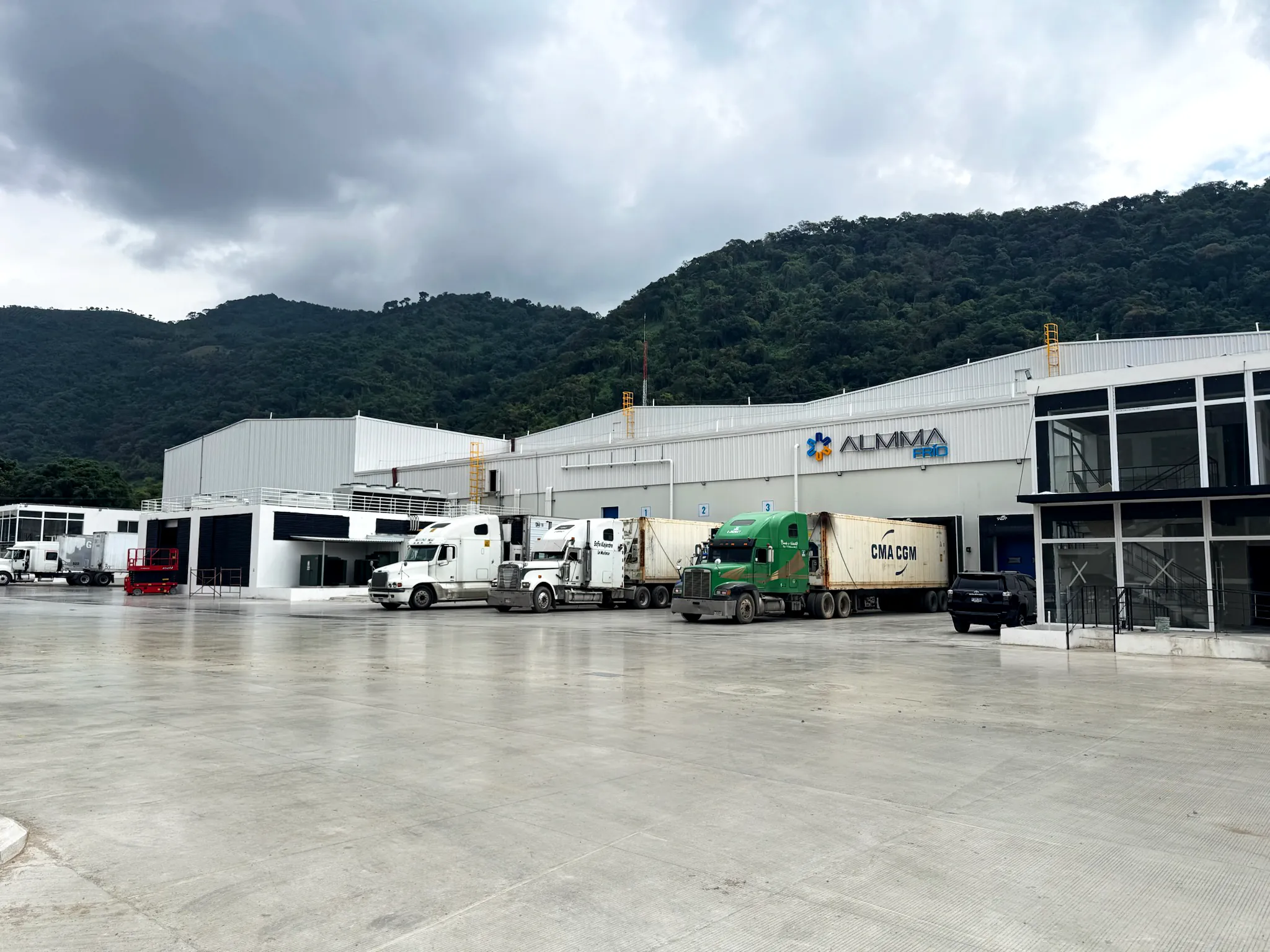 Camiones de carga estacionados junto a un almacén industrial con montañas al fondo bajo cielo nublado.
