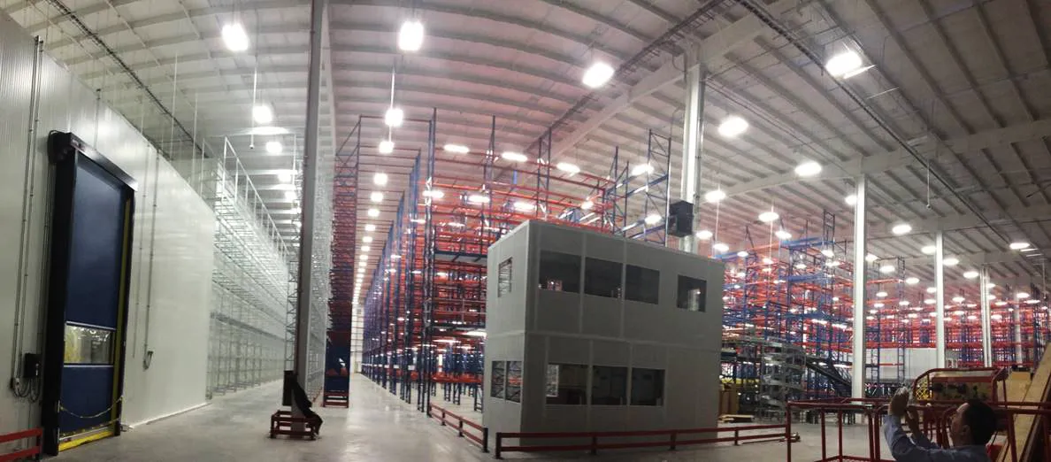 Interior amplio y iluminado de un almacén con estanterías metálicas grandes y una oficina prefabricada en el centro.