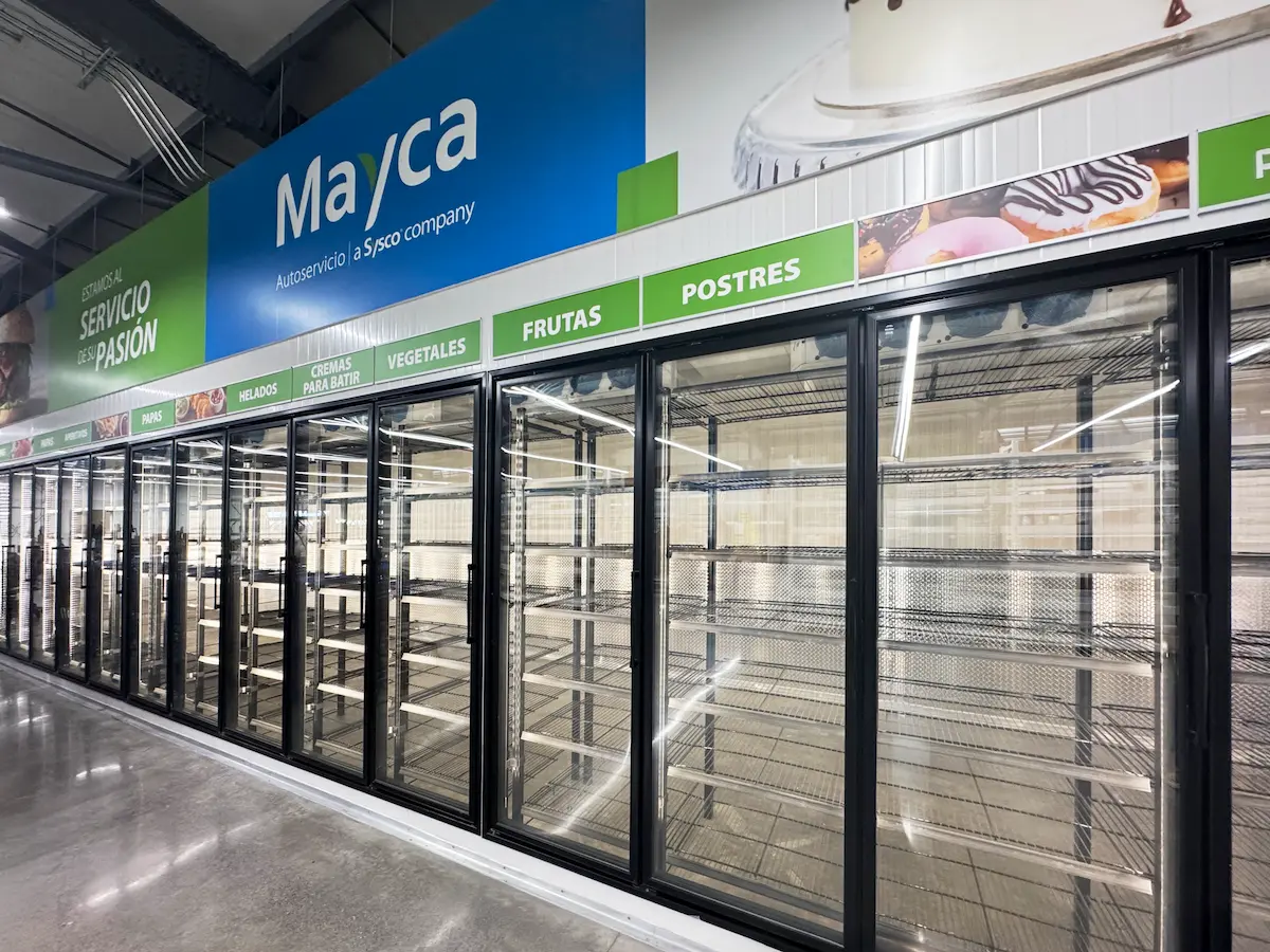 Pasillos con refrigeradores vacíos en supermercado Mayca con letreros verdes de alimentos.