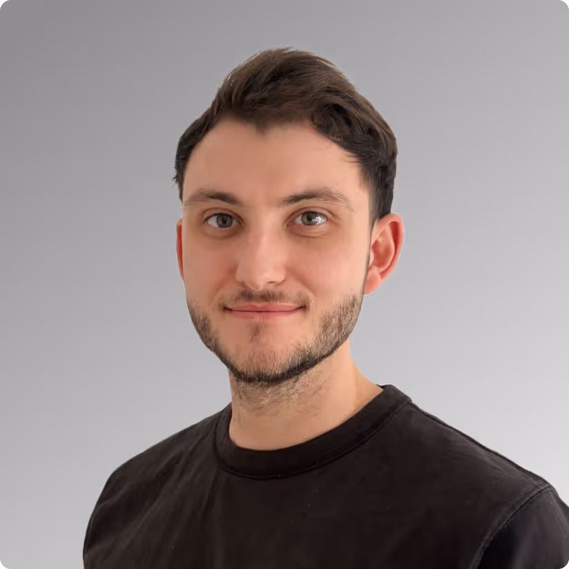 Porträt eines jungen Mannes mit kurzem braunem Haar und Bart, der ein schwarzes T-Shirt trägt vor grauem Hintergrund.