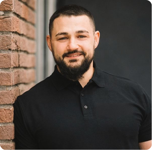 Ein Mann mit kurzem Haar und dichtem Bart lehnt an eine Backsteinwand und lächelt.