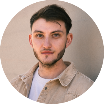 Porträt eines jungen Mannes mit dunklen Haaren und Bart, der eine beige Jacke über einem weißen T-Shirt trägt.