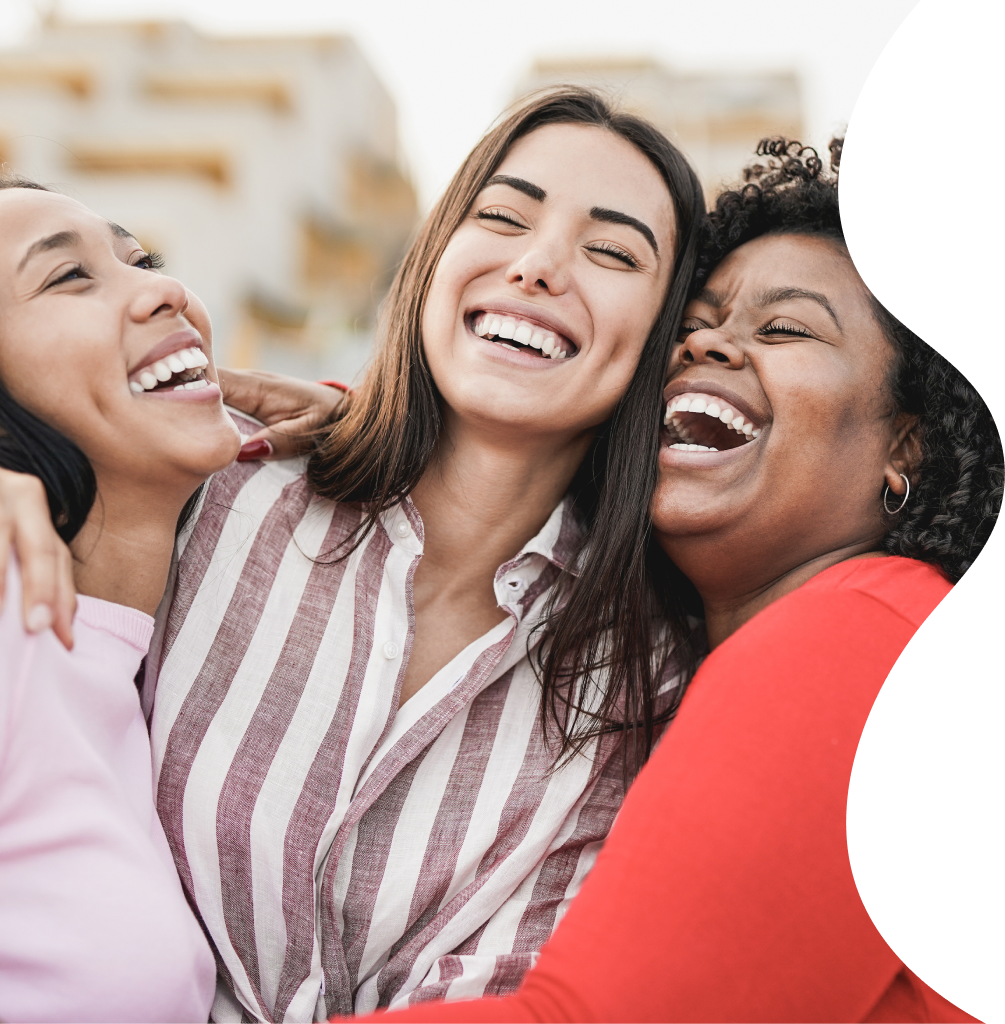 Three diverse women laughing and hugging each other outdoors.