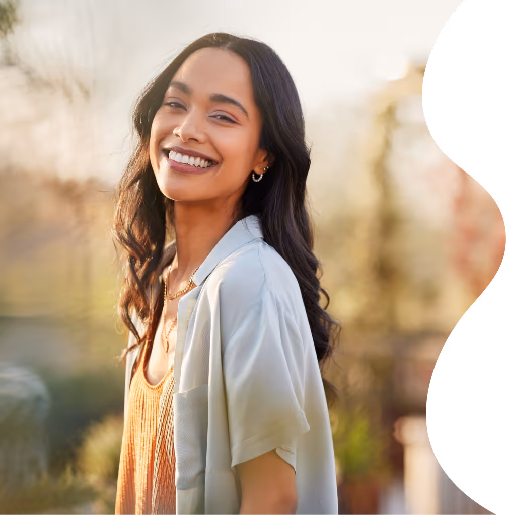 Smiling young woman with long dark hair wearing a light grey shirt over an orange top, outdoors in soft sunlight.