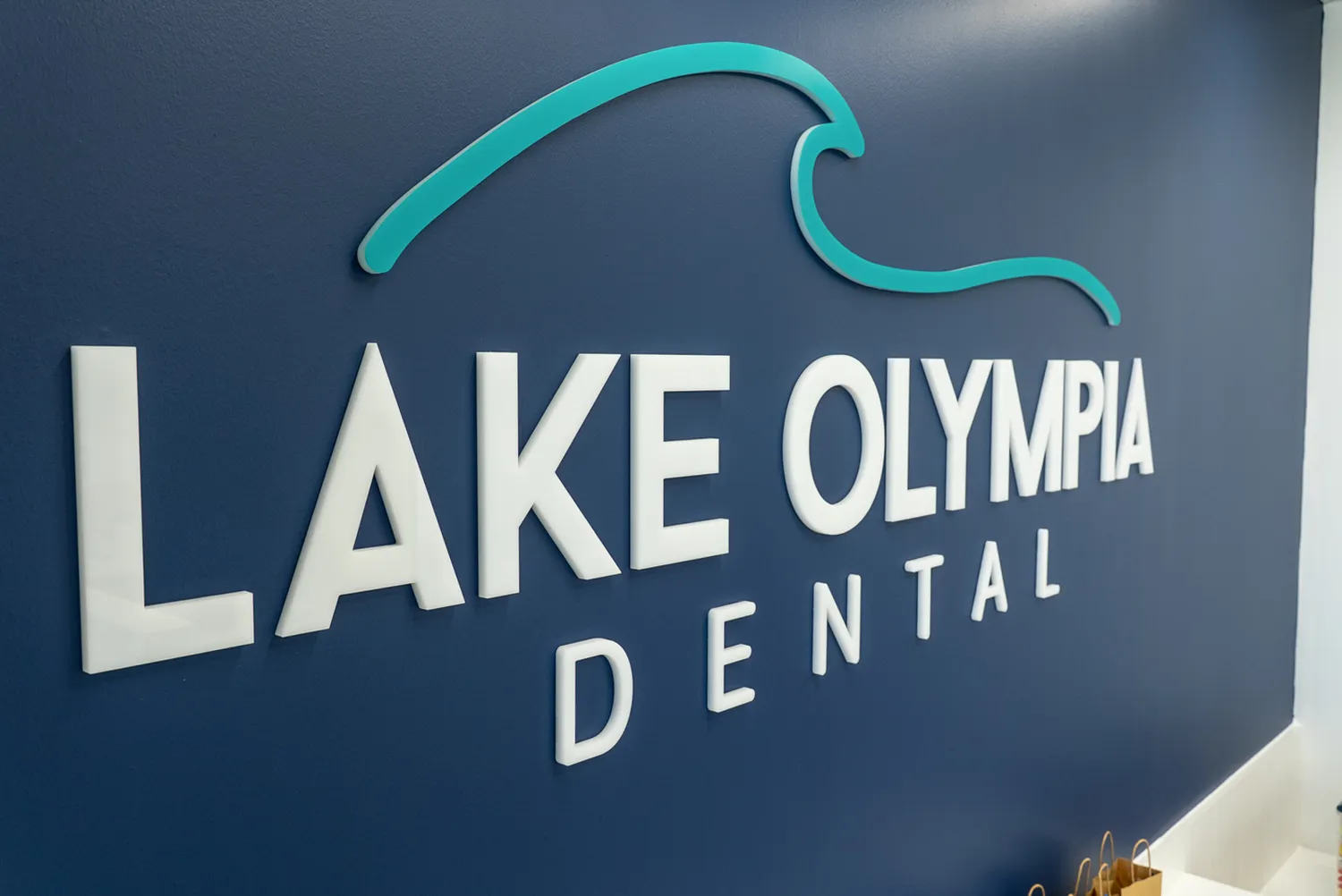 Wall sign with white letters reading 'LAKE OLYMPIA DENTAL' and a turquoise wave graphic above the text on a dark blue wall.