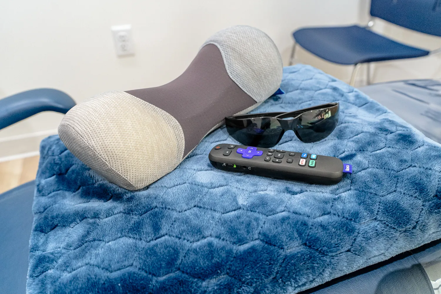 Black sunglasses, a remote control, and a gray and white neck pillow resting on a blue quilted blanket over a chair.