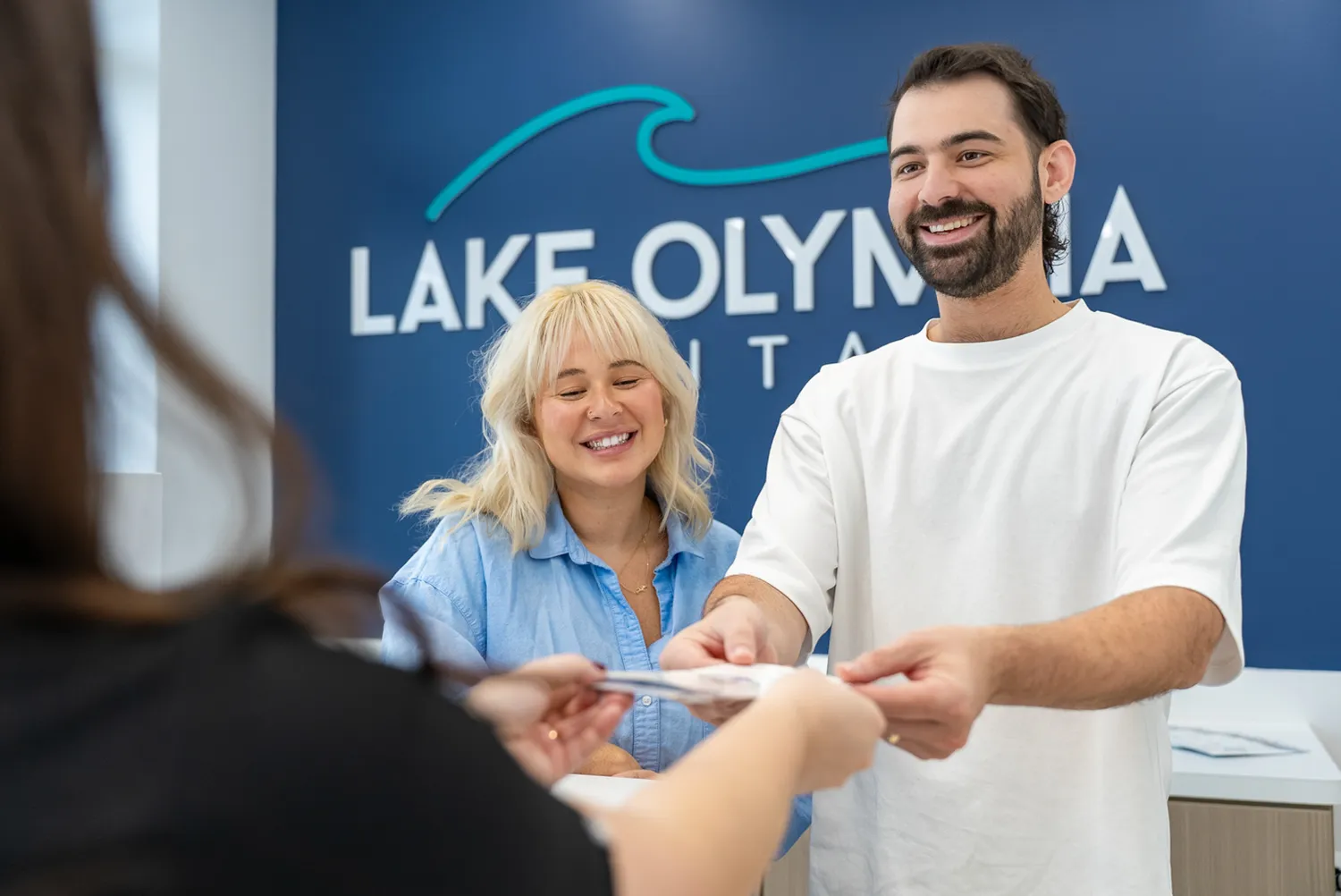 Male receptionist in white shirt handing a document to a patient with a smiling female coworker in a dental office.