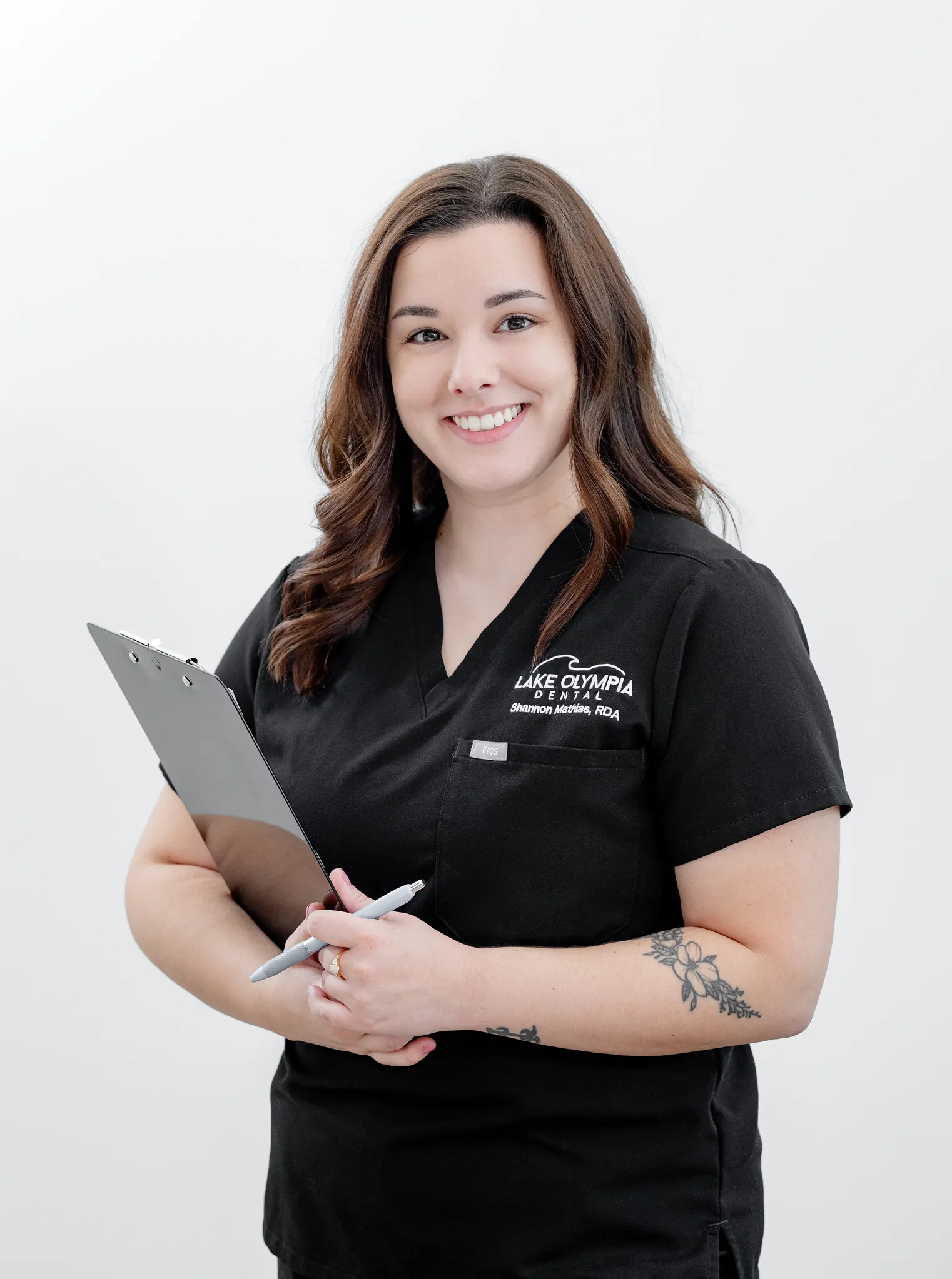 Smiling dental professional in black scrubs holding a clipboard and pen against a white background.