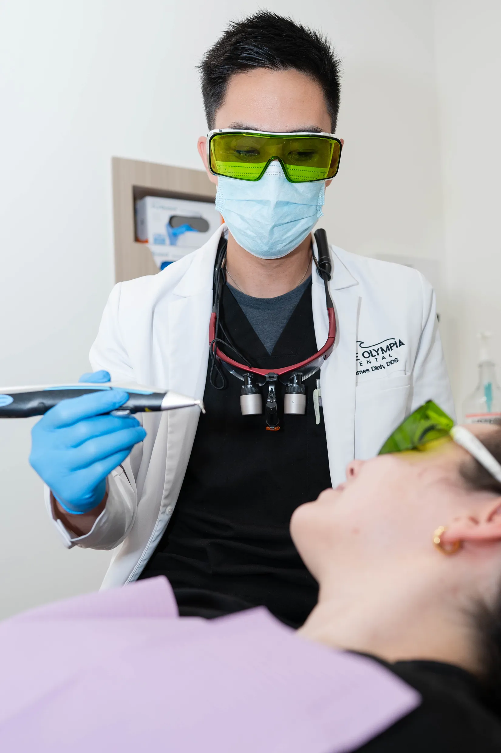 Dentist wearing mask and yellow protective glasses holding a dental laser tool near a patient.