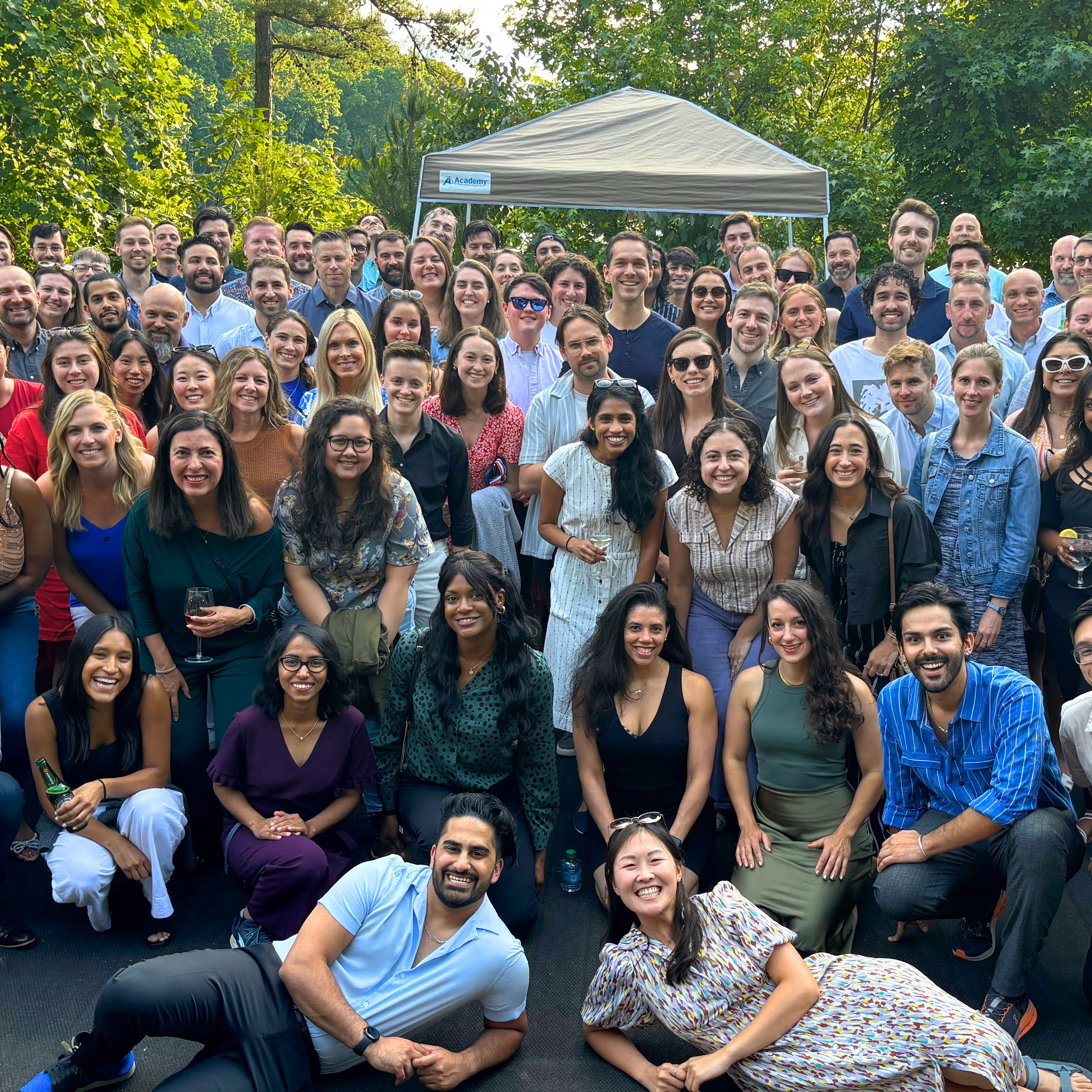 Large diverse group of people posing outdoors on a sunny day with trees and a canopy tent in the background.