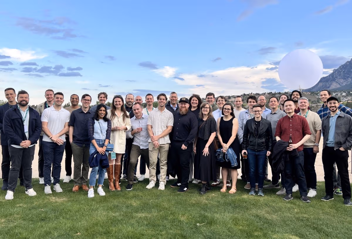 Group of diverse adults standing on grass outdoors with a mountain and large white balloon in the background.