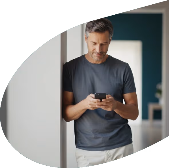 Man in gray t-shirt leaning against a wall while looking at his phone indoors.