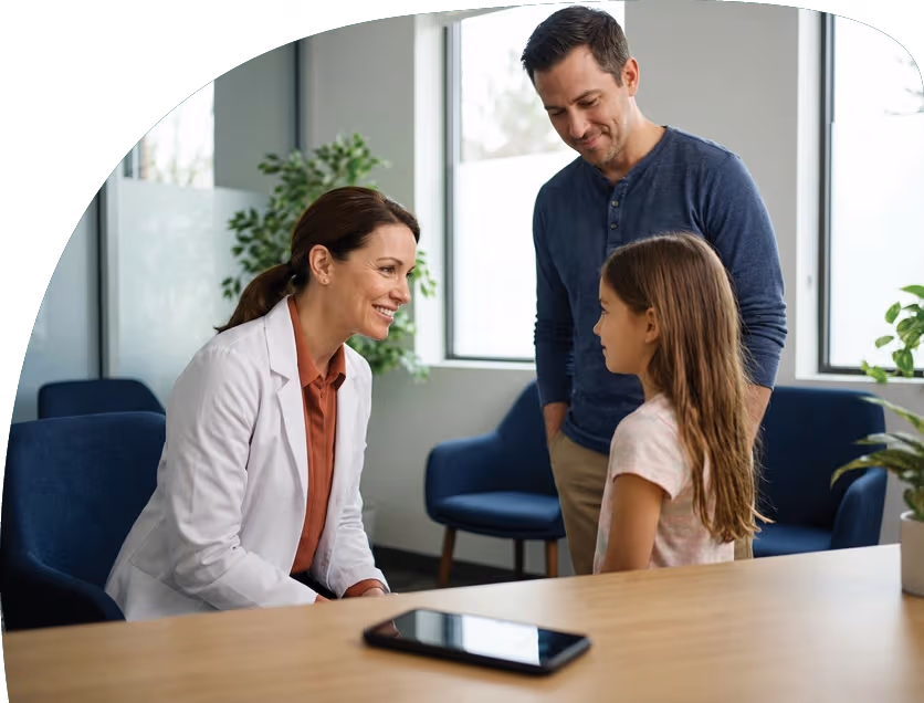 Female doctor smiling and talking to a young girl with a man standing behind her in a medical office.