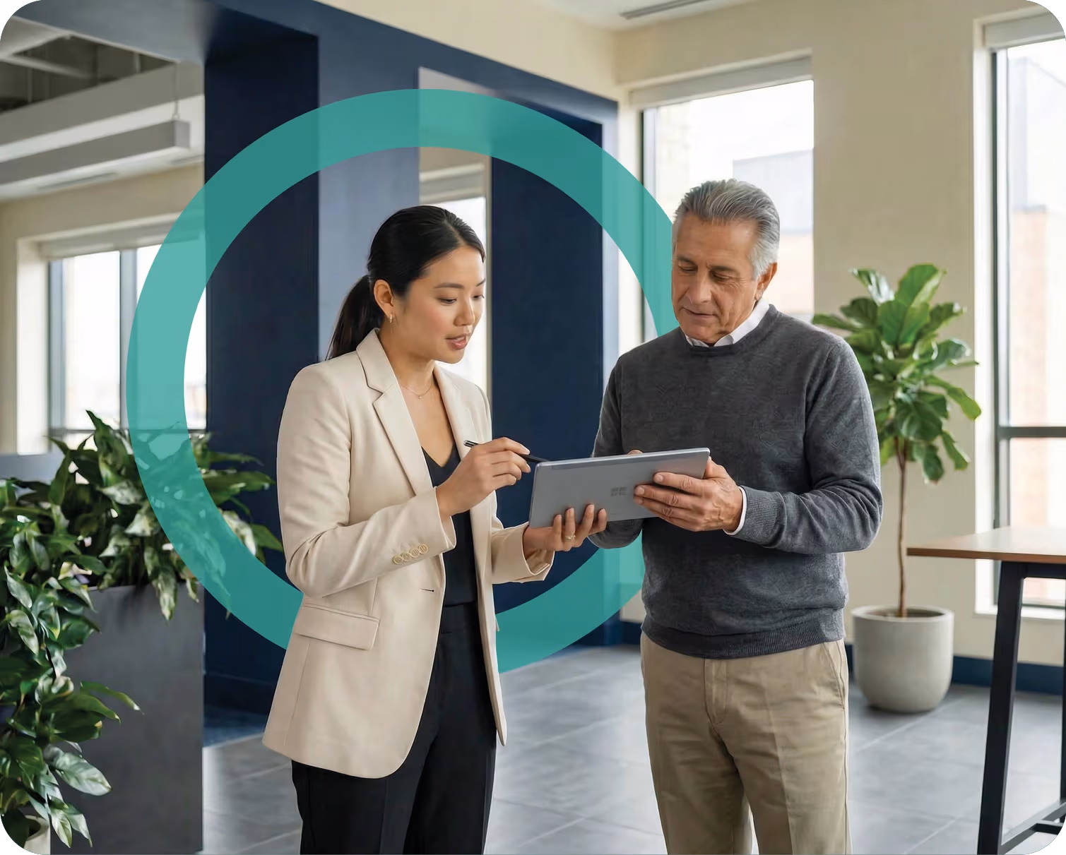Two business professionals discussing information on a tablet in a modern office with large windows and plants.