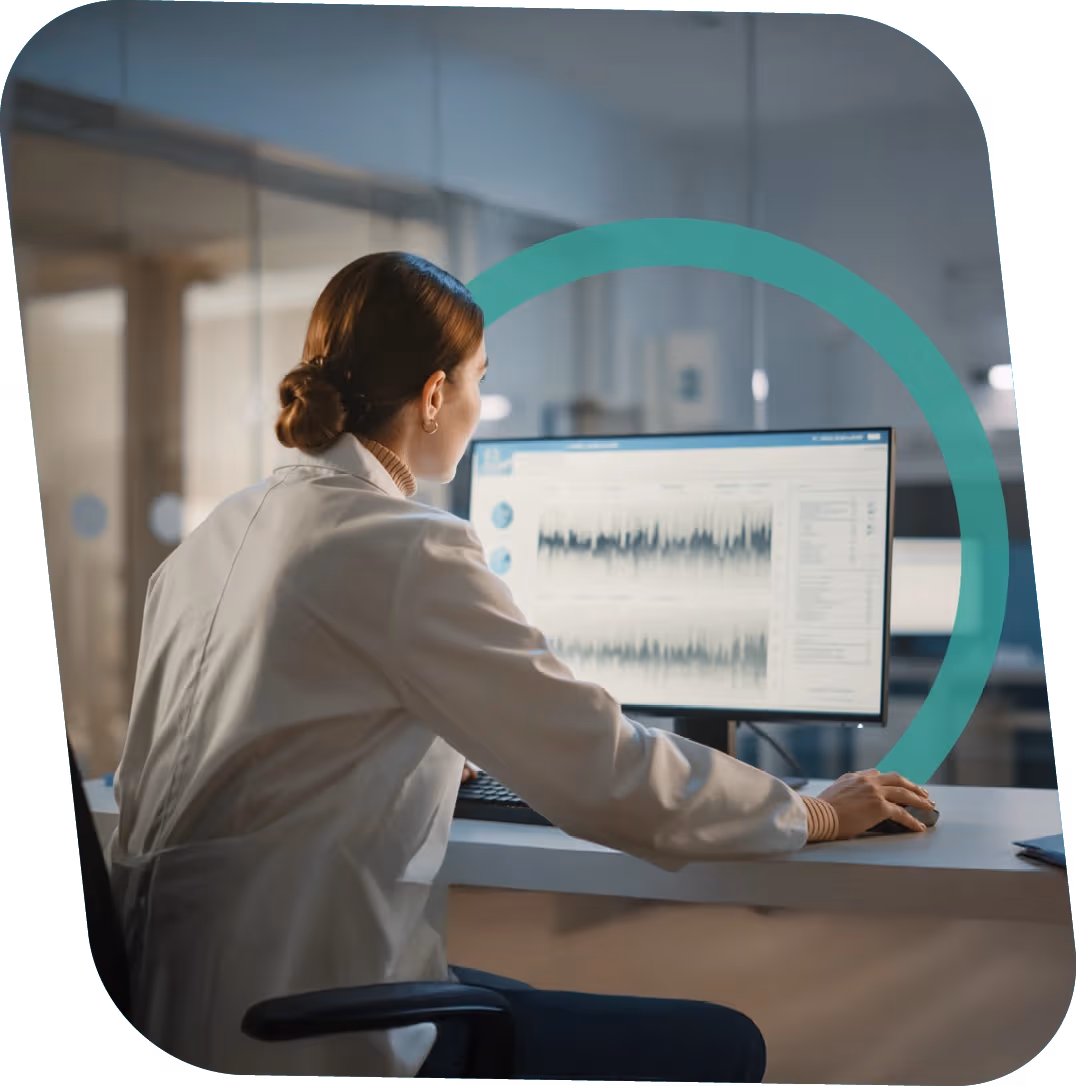 Woman in a white lab coat analyzing data charts on a computer monitor in an office setting.