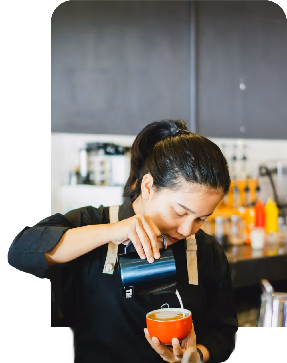 Barista in black shirt and beige apron pouring milk into a red cup to make latte art in a coffee shop.