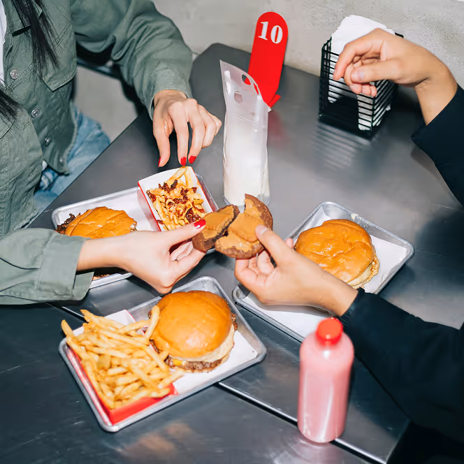 Two people sharing a broken chocolate chip cookie at a table with trays of burgers, fries, and drinks.