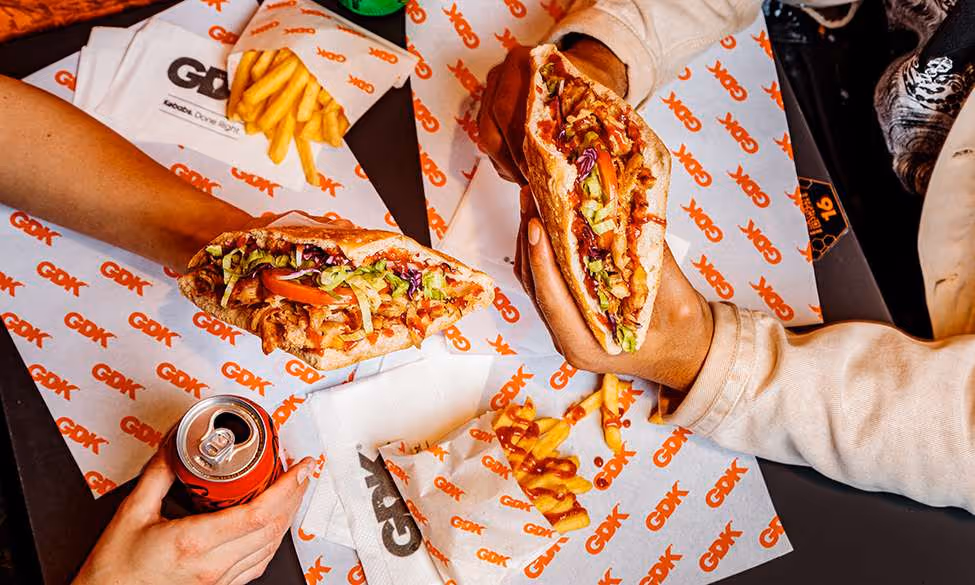 Two people holding loaded pita sandwiches with lettuce, tomato, and sauce, alongside servings of fries and a can of soda on a table with branded paper wrappers.