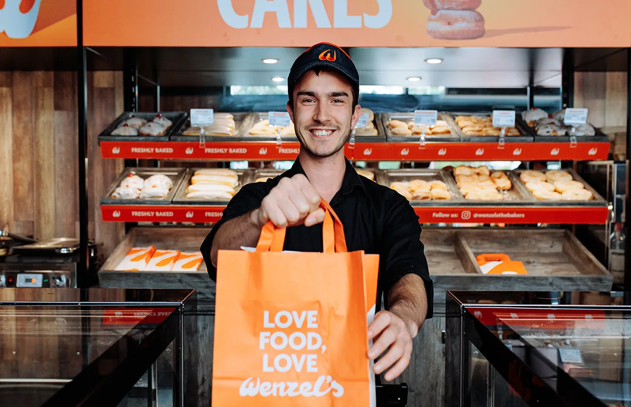 Smiling bakery employee holding out an orange Wenzel's bag with baked goods displayed in the background.