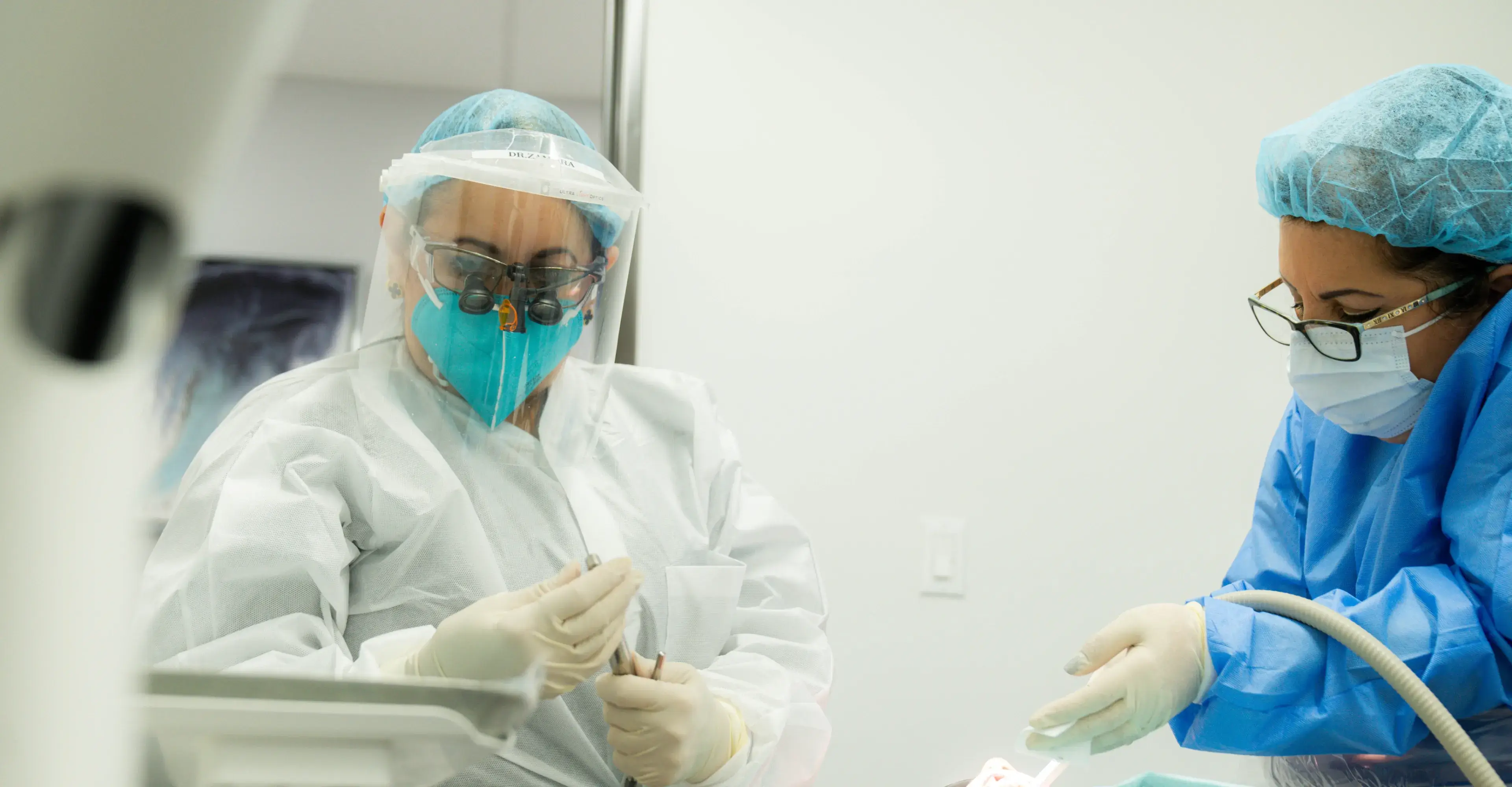 Two dental professionals wearing protective gear and masks performing a dental procedure.