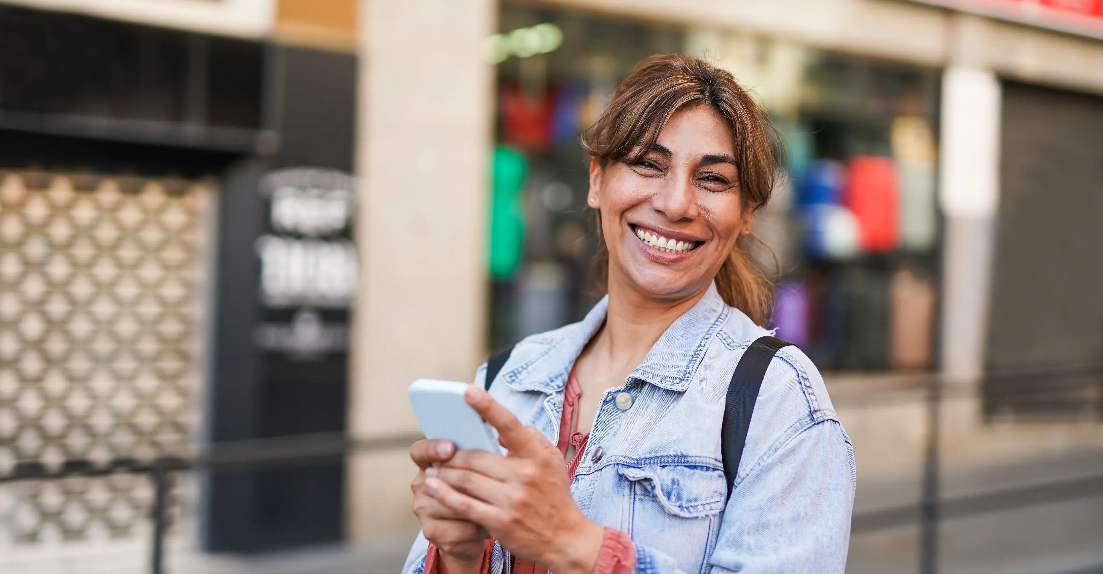 Smiling woman with light brown hair in a denim jacket holding a smartphone outdoors.