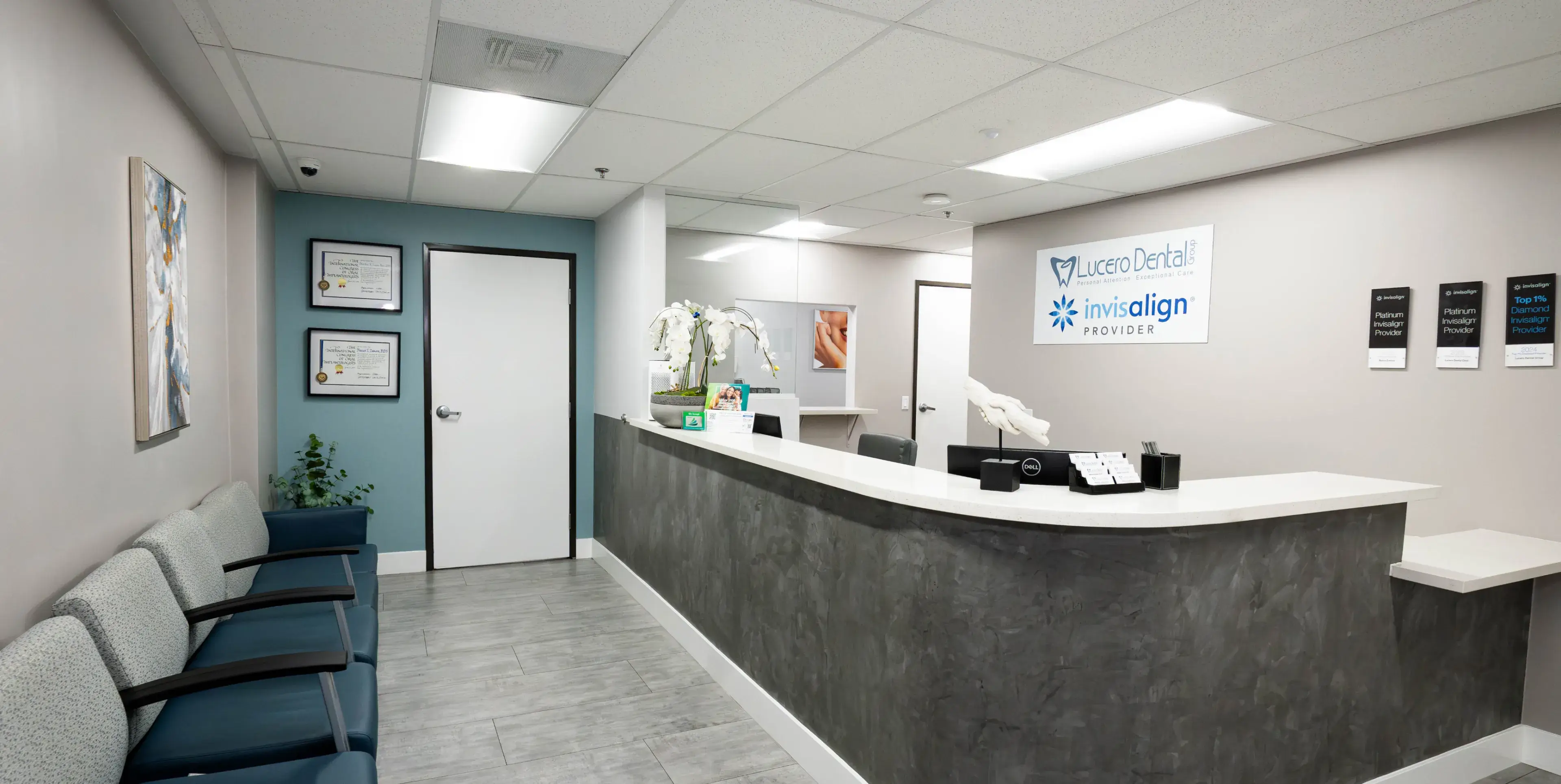Modern dental clinic reception area with gray and blue seating, reception desk, and Lucero Dental Group and Invisalign provider signs on the wall.