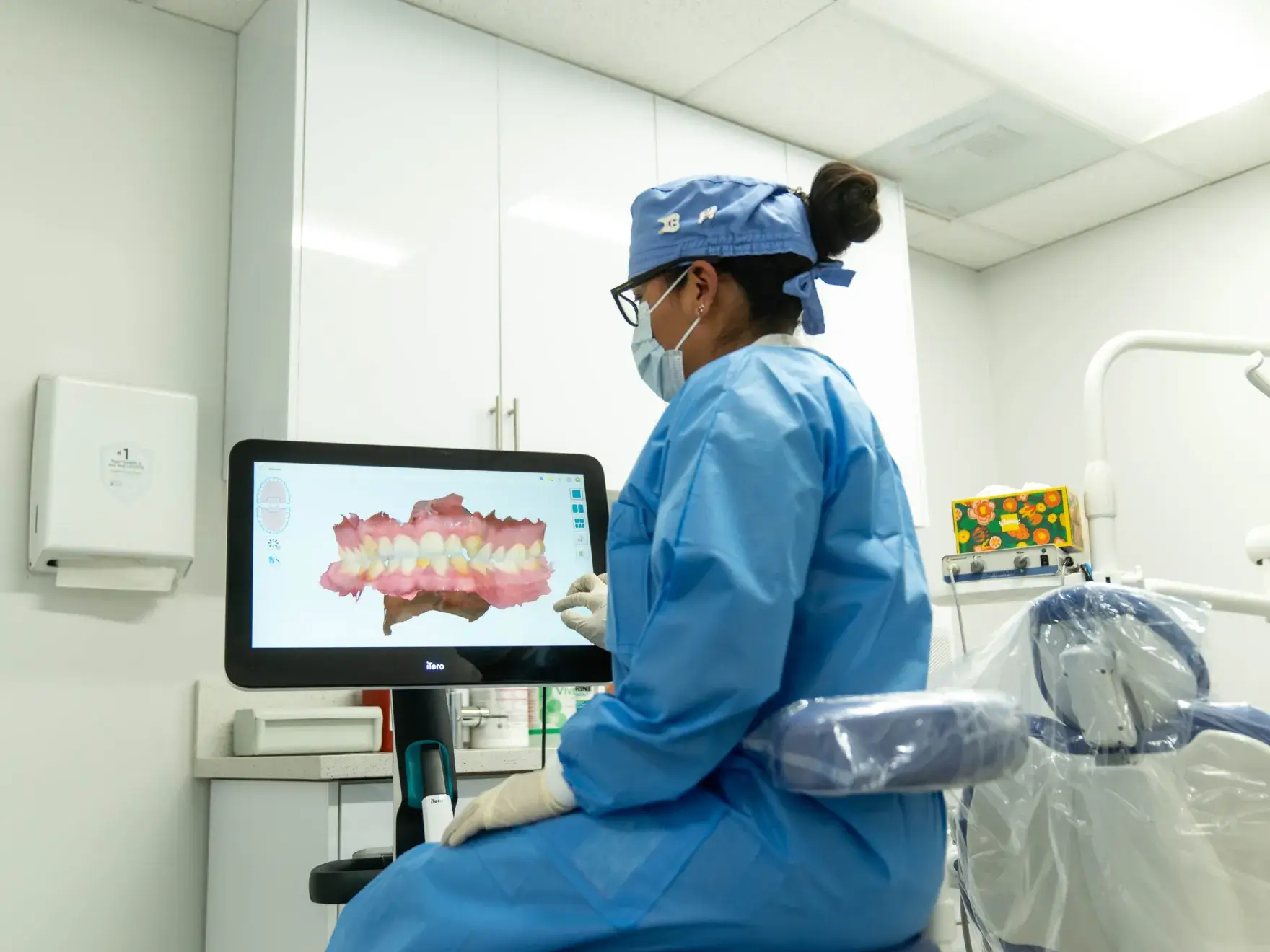 Dental professional in blue scrubs and protective gear viewing a 3D dental scan on a monitor in a dental clinic.