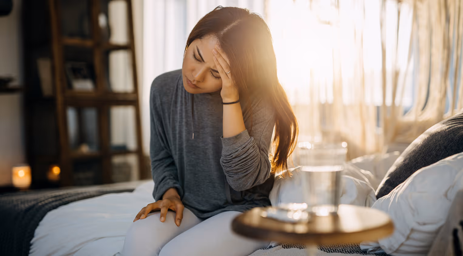 Woman sitting on a bed with her hand on her forehead, looking distressed with a glass of water on a bedside table in front of her.