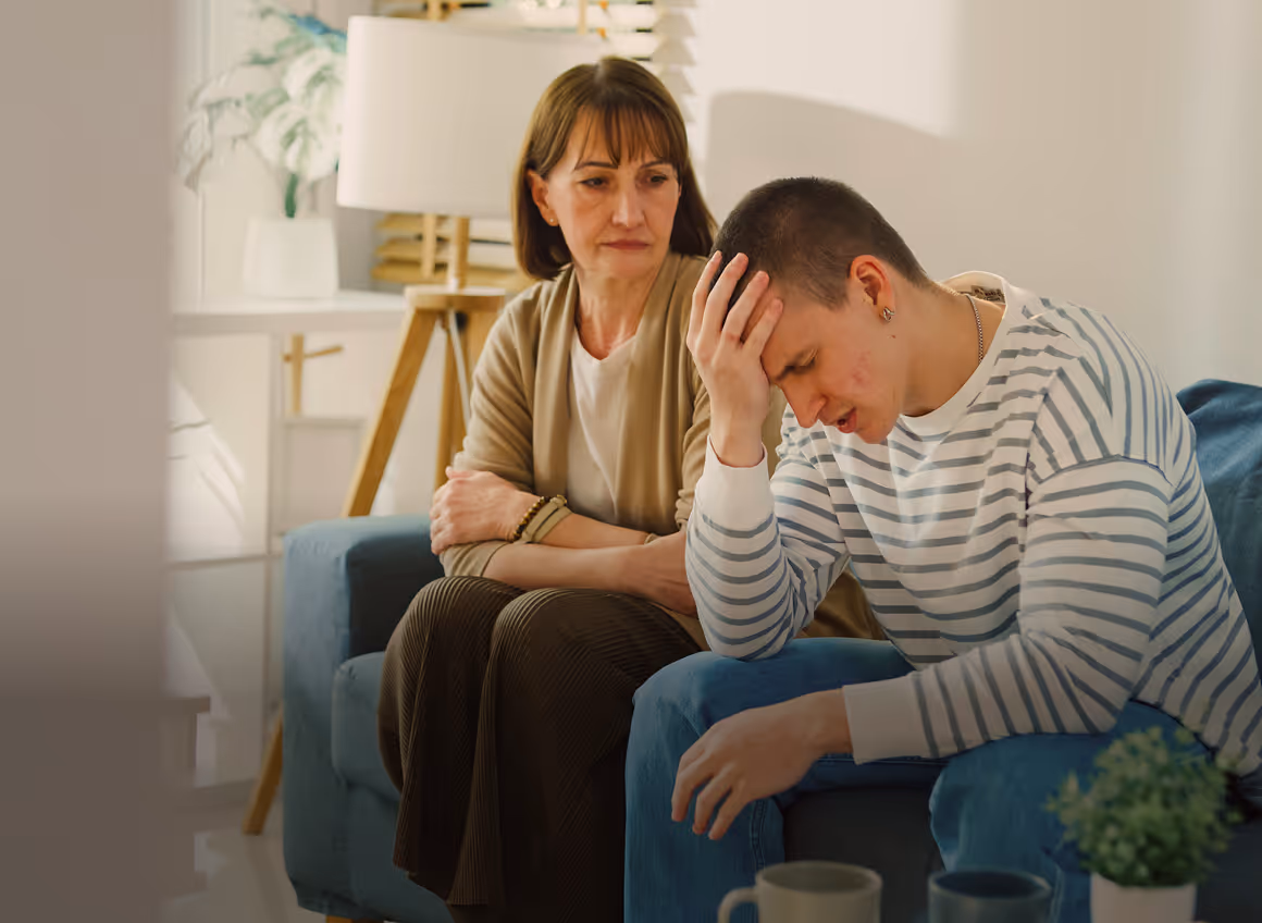 Smiling woman with curly hair affectionately hugging a teenage boy in a yellow hoodie on a couch in a cozy living room.