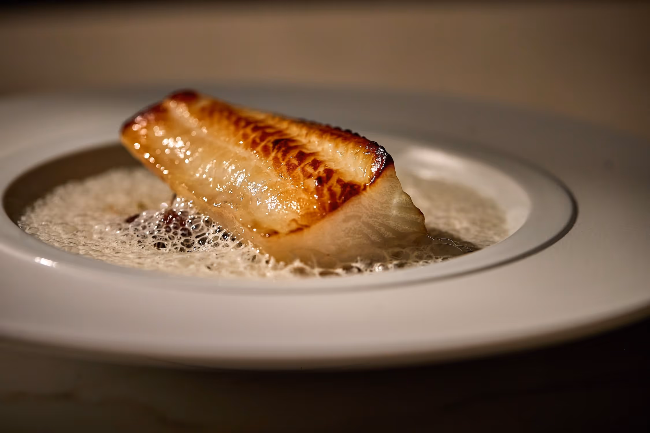 Seared fish fillet with crispy golden-brown top served in a creamy, foamy sauce on a white plate.