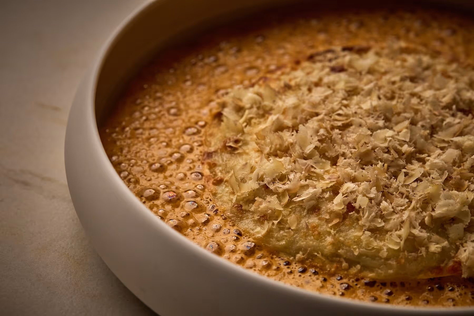 Close-up of a dish with a round food item topped with grated or shaved ingredients in a bubbling sauce.