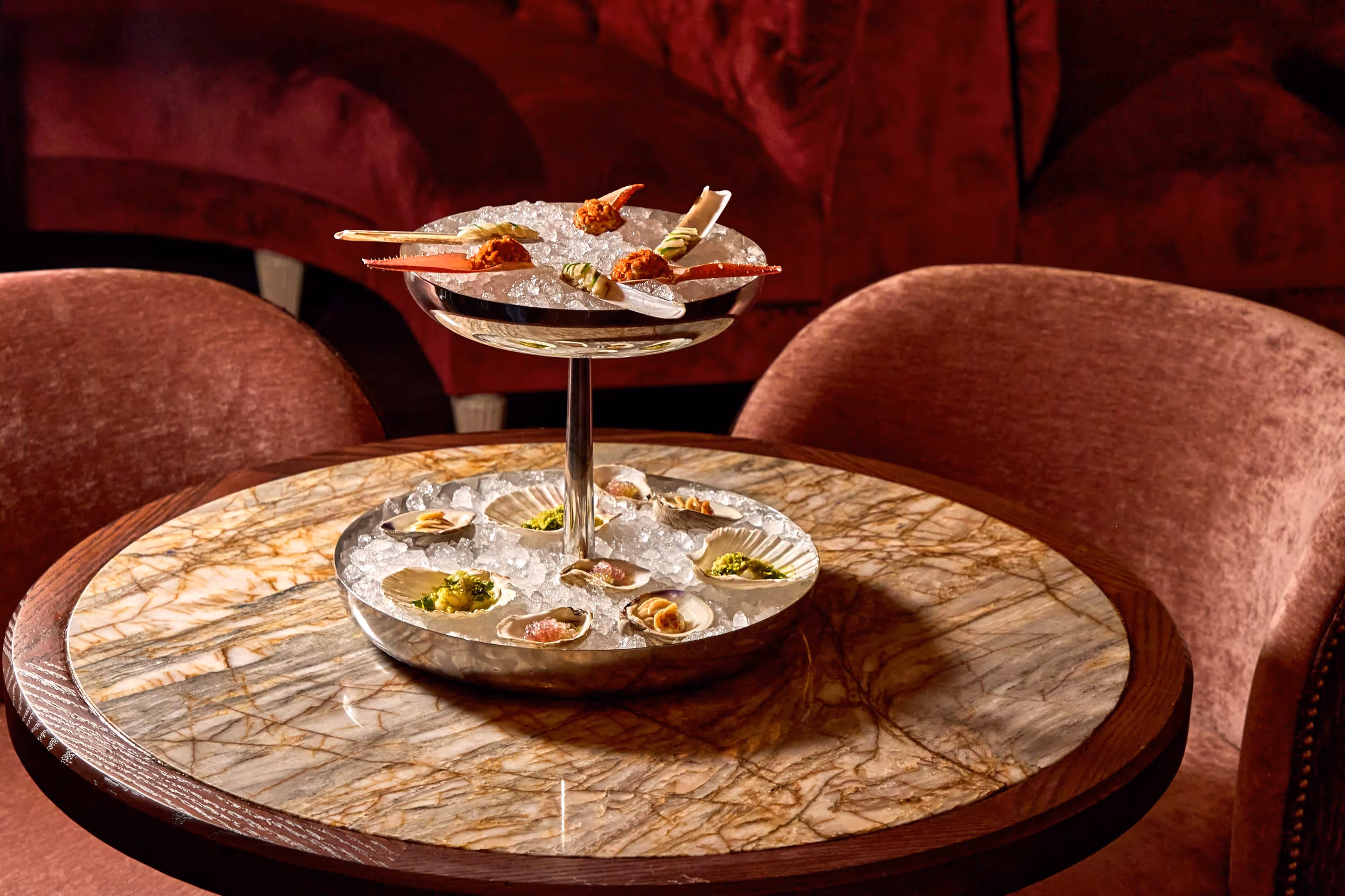 Two-tiered silver seafood platter with ice, crab claws on top tier, and various shellfish with garnishes on bottom tier, on a marble table with reddish chairs in the background.