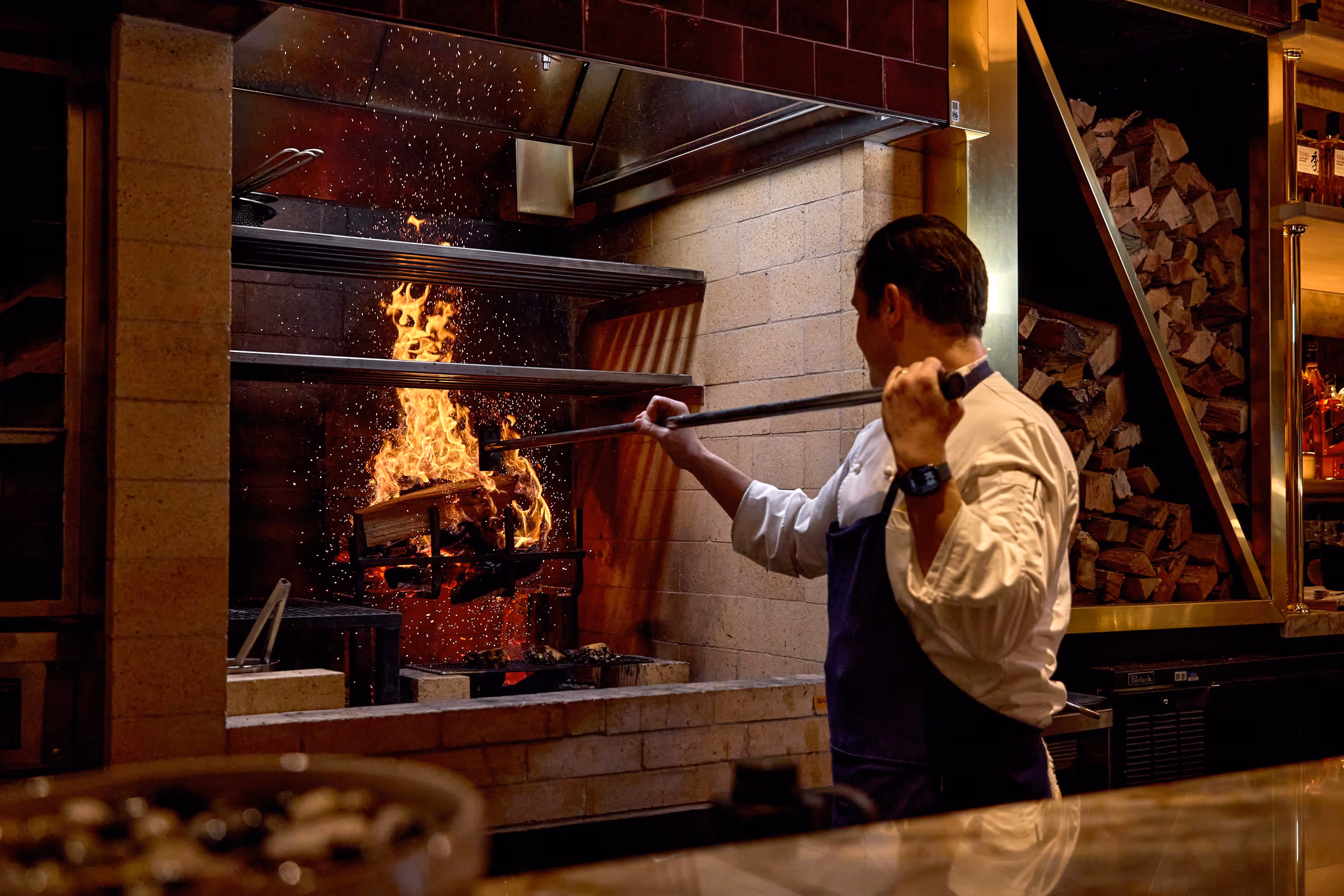 Chef tending to a wood fire in a brick fireplace with logs stacked to the side.