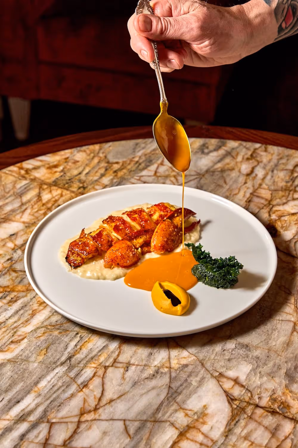 Hand pouring orange sauce from a spoon onto a plated gourmet lobster dish with a garnish and puree on a marble table.