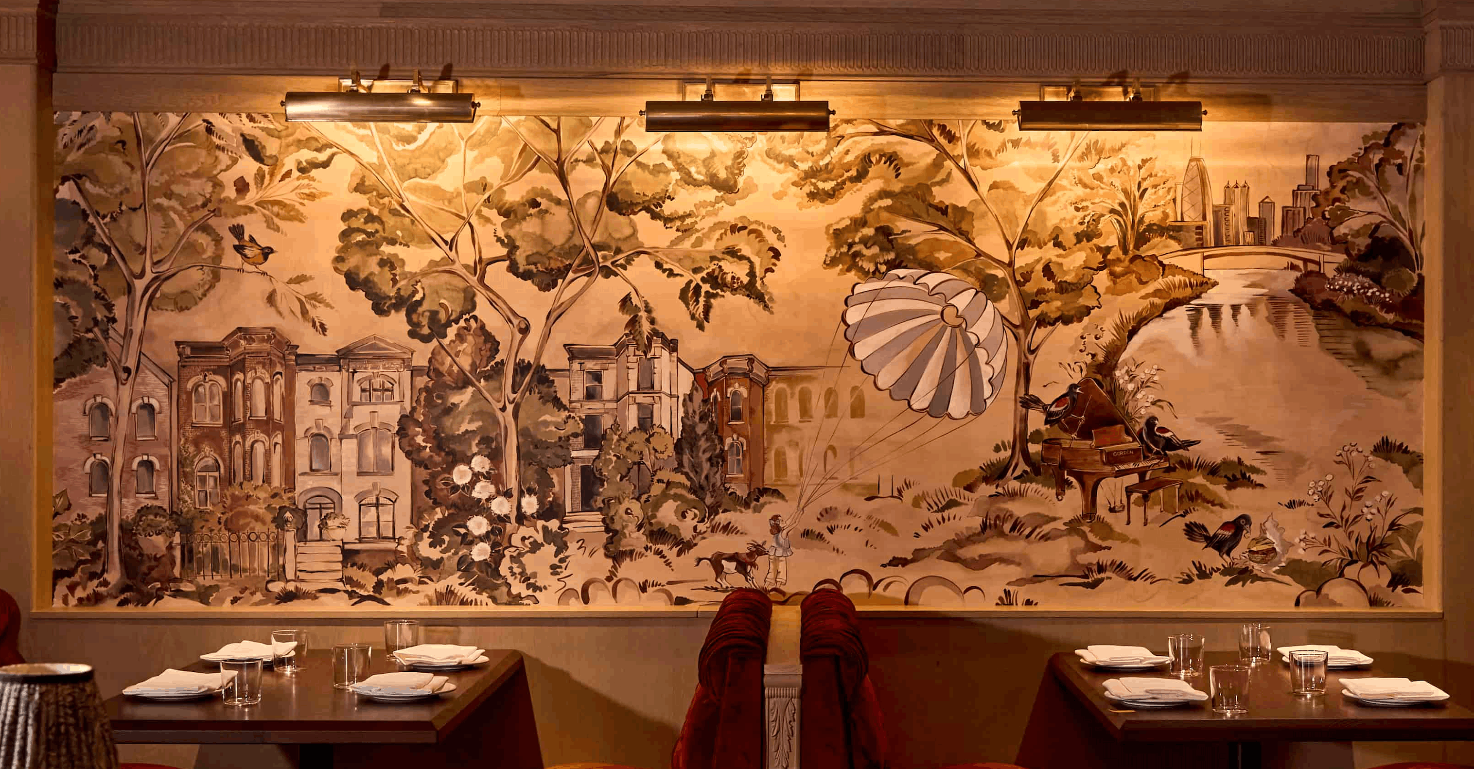 A large sepia-toned mural covering the restaurant wall, depicting a sweeping landscape with Victorian buildings, lush trees, a hot air balloon, a grand piano, birds, and a city skyline in the distance, with set dining tables in the foreground.